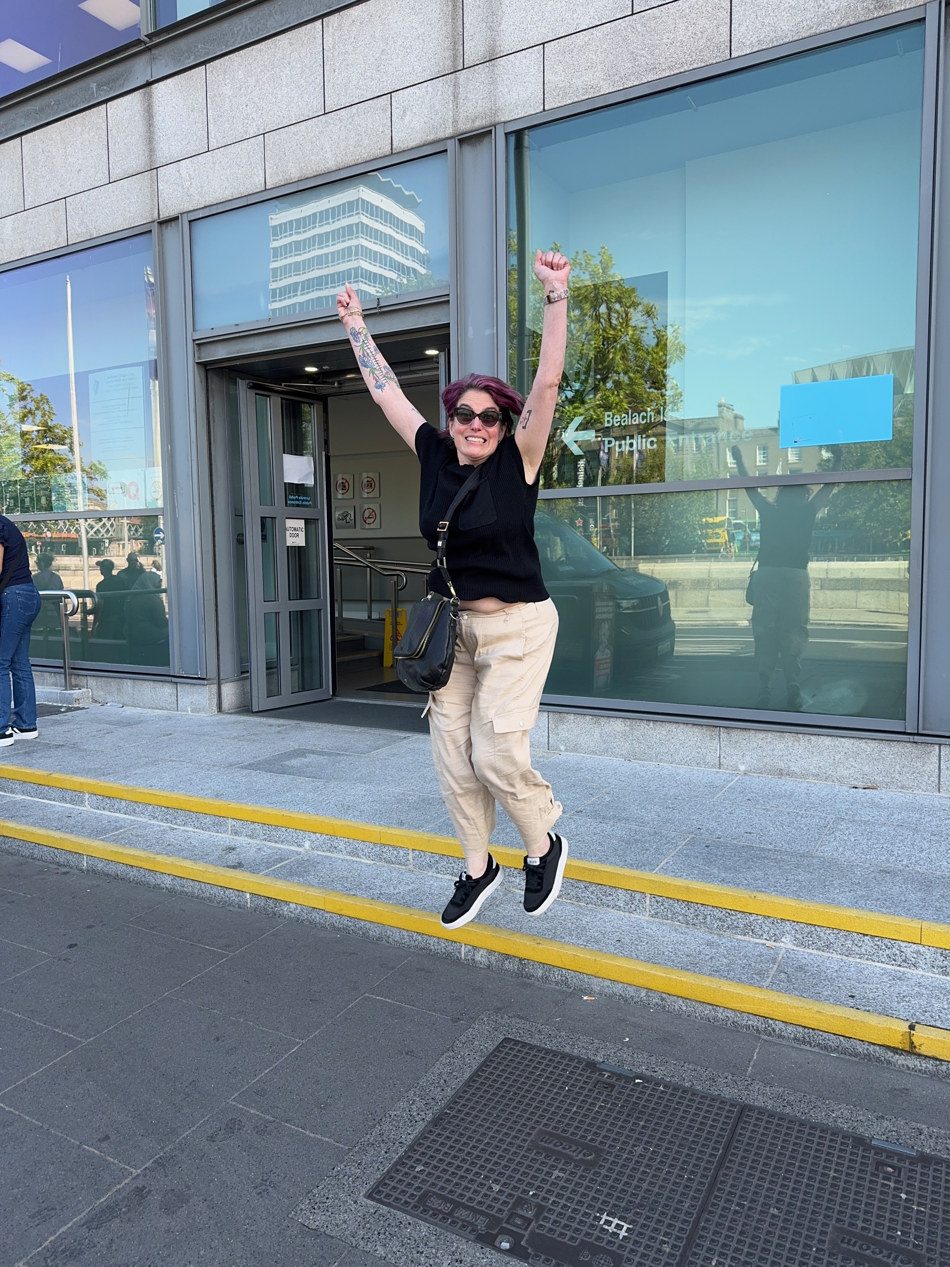 Image of Megan jumping for joy after getting the stamp 4 on her passport. She is wearing tan pants with a black short-sleeved shirt and a bag across her body. She is wearing sunglasses and her arms are in the air and feet are off the ground.