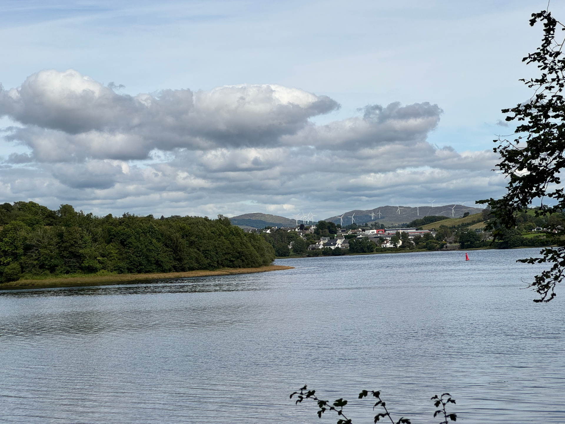 Image of the entry to the Atlantic Ocean as seen from the Mac Manus Estate in Donegal. across water, to the left, are a copse of trees. In the middle are homes with a mountain range in the background. Windmills can be seen along the mountains.